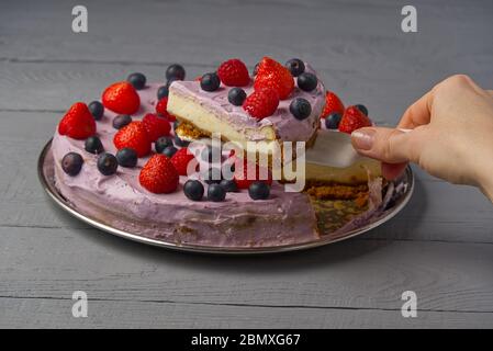 La femme a divisé le cheesecake et prend une tranche pour l'offrir. Cheesecake maison avec fraise, myrtille et framboise sur fond de bois gris. Banque D'Images