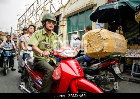 Un vieil homme habillé de vêtements militaires fait une moto rouge dans les rues animées d'un marché local à Hanoi Old Quarter, Vietnam. Son vélo Banque D'Images