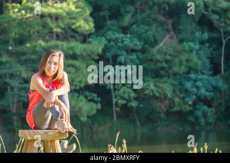 Portrait Asian femme assise sur chaise en bois fond flou eau et arbres. Banque D'Images