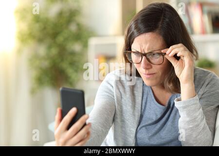 Femme adulte ayant des problèmes de vue portant des lunettes de vue lisant sur un smartphone assis sur un canapé à la maison Banque D'Images