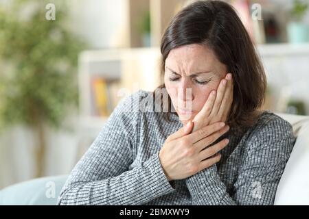 Femme d'âge moyen en douleur souffrant de maux de dents touchant le visage assis sur un canapé à la maison Banque D'Images