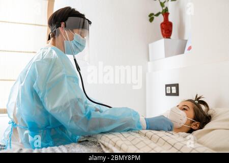 Médecin protégé par un équipement de protection personnelle examinant une petite fille avec un stéthoscope à la maison. Banque D'Images