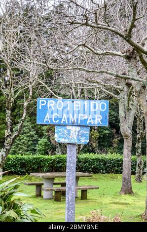 Vieux signe bleu avec texte Proibido Acampar en langue portugaise. TRADUCTION: Camping interdit. Parc avec arbres verts, bancs et tables en arrière-plan. Pas de panneau de camping, pas de séjour de nuit. Banque D'Images