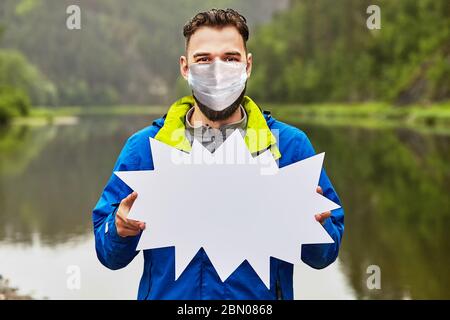 Un homme barbu dans un masque chirurgical montre une forme de publicité sculptée contre un paysage naturel. Banque D'Images