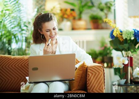 femme moderne souriante d'âge moyen dans un chemisier blanc et une veste avec ordinateur portable appliquant le rouge à lèvres tout en étant assise sur un canapé à la maison moderne par beau temps. Banque D'Images