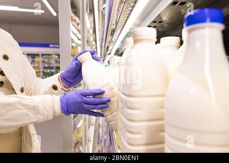 Gros plan des mains de femme en gants médicaux choisit les produits laitiers en ouvrant le réfrigérateur dans le supermarché. Mesures de protection contre la pandémie du coronavirus, co Banque D'Images