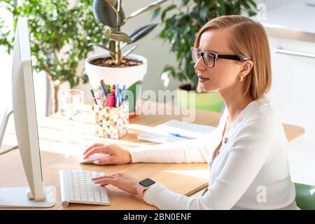 Femme pigiste pensive dans des lunettes travaillant sur l'ordinateur à partir du bureau à domicile pendant la quarantaine en raison du coronavirus. Espace de travail confortable entouré de plantes. Activé Banque D'Images