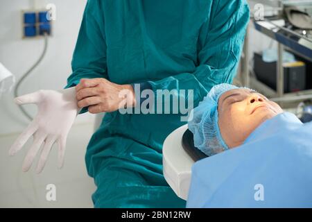 Femme sénior couché sur une table d'opération en clinique d'ophtalmologie avec ses yeux fermés, chirurgien mettant des gants en caoutchouc Banque D'Images