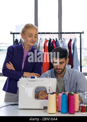 Les créateurs de mode senior contrôlent étroitement les nouveaux concepteurs dans leur travail qui nécessite des machines à coudre électriques. Homme et femme travaillant à la maison cl Banque D'Images