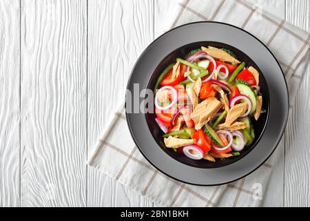 Salade de légumes avec thon en conserve : tomates, concombre, oignon rouge et haricots verts servis sur une assiette noire sur fond de bois blanc, en haut vie Banque D'Images