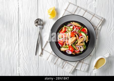 Salade de thon avec sauce au citron et légumes : tomates, concombre, oignon rouge et haricots verts servis sur une assiette noire sur fond de bois blanc, fla Banque D'Images