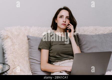 La fille travaille à la maison et a pensé à quelque chose. Portrait d'une femme parlant sur un téléphone portable devant son ordinateur portable à la maison. Banque D'Images