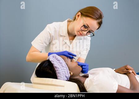 Le médecin cosmetologue fait la procédure d'augmentation de lèvre d'une belle femme africaine dans un salon de beauté. Cosmétologie soins de la peau. Banque D'Images