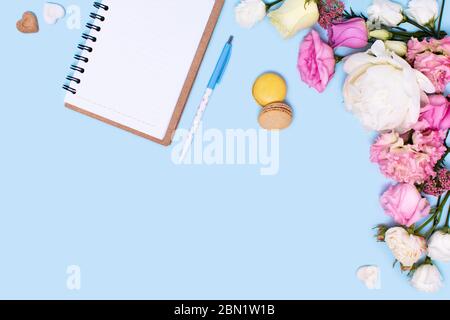 Carte de vœux ou invitation à un congé ou une fête. Roses et pivoines, bloc-notes, stylo et gâteaux macaroni avec des sucreries sur fond bleu. Banque D'Images
