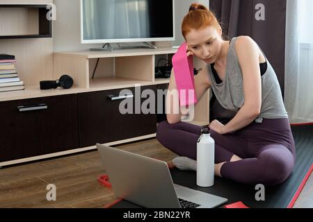 La fille aux cheveux rouges est assise à la maison dans un uniforme sportif avec une bande élastique dans sa main et regarde avec une grande déperance l'ordinateur portable. Banque D'Images