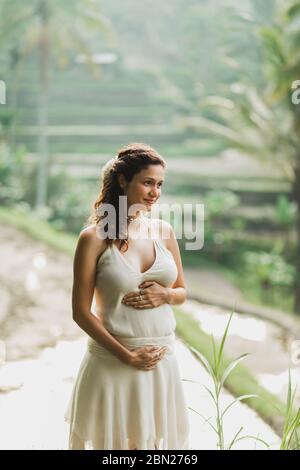 Jeune femme enceinte en robe blanche avec vue sur les rizières en terrasses de Bali dans la lumière du soleil du matin. Harmonie avec la nature. Concept de la grossesse. Banque D'Images