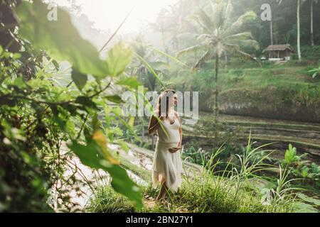 Jeune femme enceinte en robe blanche avec vue sur les rizières en terrasses de Bali dans la lumière du soleil du matin. Harmonie avec la nature. Concept de la grossesse. Banque D'Images