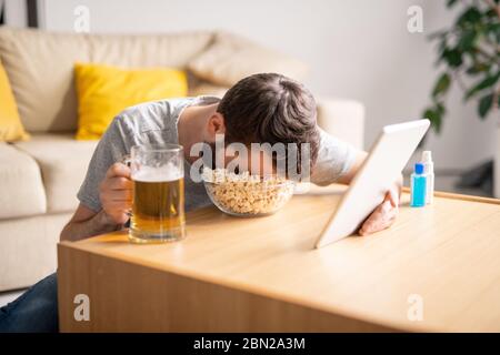 Homme fatigué assis à une table basse et dormant dans un bol de pop-corn tout en regardant la vidéo sur une tablette à la maison, concept de période d'isolement Banque D'Images