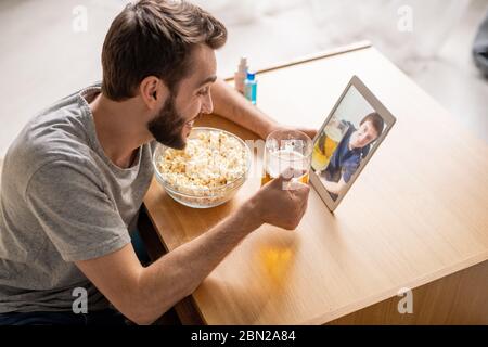 Un jeune homme positif et enthousiaste boit de la bière avec un ami et mange du pop-corn tout en utilisant le chat vidéo sur une tablette Banque D'Images