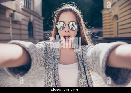 Une fille choquée dans des lunettes de soleil et la bouche ouverte fait le selfie sur un appareil photo. Elle porte une tenue décontractée, marchant à l'extérieur de la ville Banque D'Images