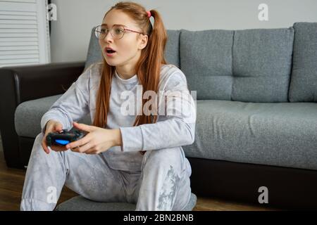 Fille rouge avec queues dans des verres est assis sur le sol et joue avec enthousiasme joystick. Banque D'Images