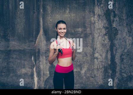 Excellent travail! Bonne jeune femme sportive dans une tenue d'entraînement élégante avec corde de saut sur ses épaules est souriante, debout dehors sur un mur de béton Banque D'Images