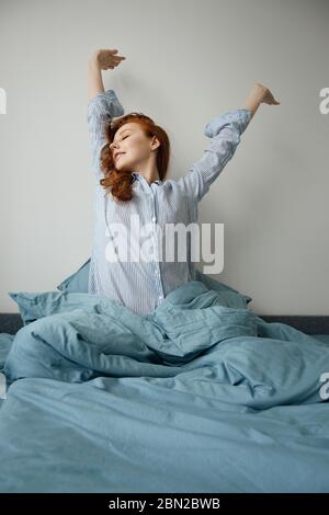 La fillette aux cheveux rouges en pyjama rayé se place dans le lit et tire les mains vers le haut. Banque D'Images