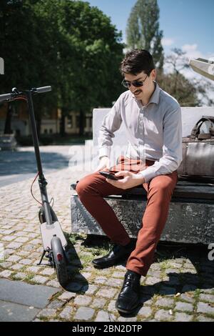 Homme souriant avec téléphone et scooter assis sur un banc. Jeune homme avec moustache et lunettes de soleil assis sur un banc à côté du scooter dans le centre-ville. Banque D'Images