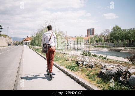 Dos de l'homme sur un scooter électrique conduisant sur la route au bord de la rivière. Vue arrière de l'homme d'affaires portant une chemise et un sac en conduisant le scooter sur le trottoir. Banque D'Images