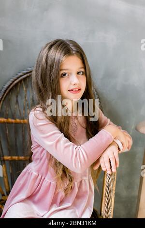 Jolie petite fille dans la belle robe rose est assis sur la chaise en bois et regarde rêveur, beauté et mode concept, portrait d'intérieur Banque D'Images