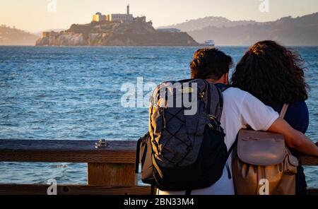 Un jeune couple debout sur le rail sur l'embarcadère 39, la promenade des pêcheurs à San Francisco, qui contemple la baie jusqu'à la prison d'Alcatraz au coucher du soleil. Banque D'Images