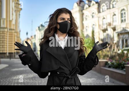 Charmante femme brune dans un masque de protection noir avec des gants de latex se tient sur la rue vide ensoleillée de la ville pendant la quarantaine de COVID-19. Elle porte un noir Banque D'Images