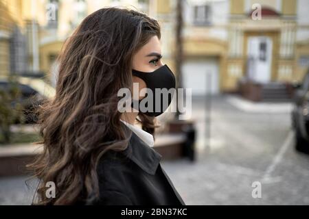Magnifique femme brune dans un masque de protection noir se tient sur vide rue ensoleillée de la ville pendant la quarantaine de COVID-19. Elle porte un trench-coat noir avec un Banque D'Images