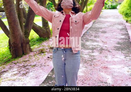 Une jeune femme vêtue de rose portant un masque facial en tissu cousu de protection à la maison jouant dans les pétales de fleur de cerisier tombés Banque D'Images