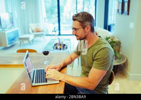 Homme travaillant à la maison à l'ordinateur portable, buvant du vin rouge dans la cuisine Banque D'Images
