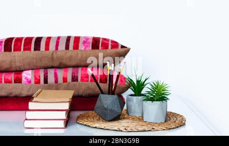 Décoration intérieure chaleureuse : crayons dans le support, pile de livres, plantes en pots sur un support en osier, oreillers et écossais sur une table blanche. Distance home éduca Banque D'Images