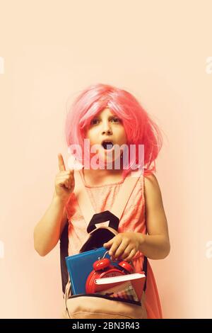 Enfant avec réveil et livres. Fille avec perruque en robe rose avec sac à dos. Retour à l'école et concept d'apprentissage. L'élève a une idée, isolée sur fond rose clair. Banque D'Images