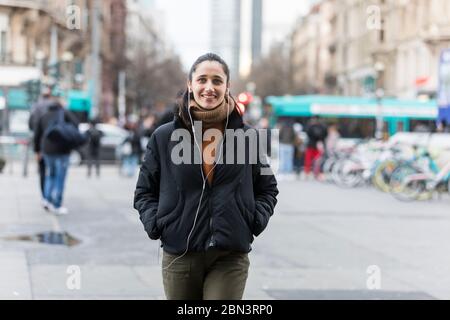 Jeune femme indienne marchant dans la rue en écoutant des écouteurs et en souriant à l'appareil photo. Banque D'Images