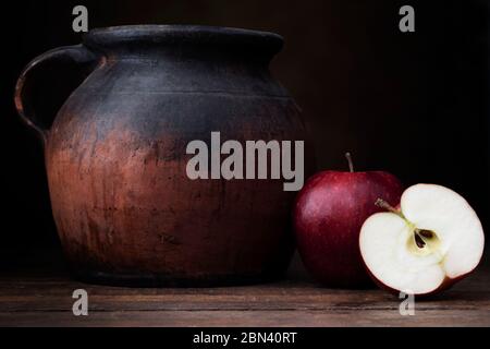 Deux pommes rouges, une toute une coupée, avec une vieille verseuse en céramique. Prise de vue au niveau des yeux avec une sensation rustique sombre. Banque D'Images