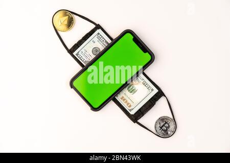 Téléphone avec écran vert sur un masque fait de 100 dollar de bancnote et bitcoin, pièces éthérées Banque D'Images