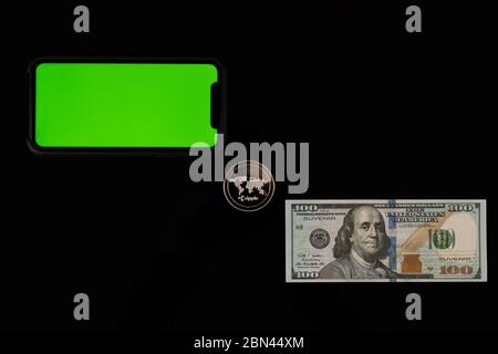 Pièce de monnaie ondulée entre billet de banque et téléphone avec écran vert isolé sur noir Banque D'Images