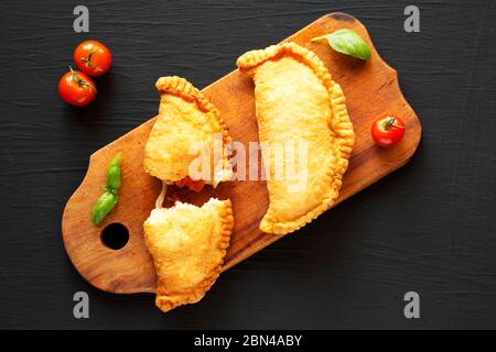 Calzone de panzerotti italien maison, accompagné de tomates et de mozzarella, sur un panneau rustique en bois sur fond noir, vue du dessus. Pose à plat, au-dessus Banque D'Images