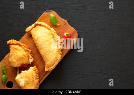 Calzone de panzerotti italien maison, accompagné de tomates et de mozzarella, sur un panneau rustique en bois sur fond noir, vue du dessus. Pose à plat, au-dessus Banque D'Images