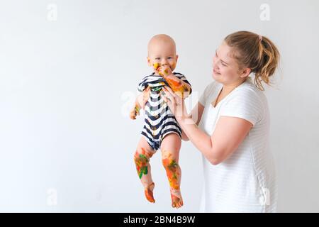 Mère tient joyeux craie bébé sale jouant avec de la peinture sur fond blanc de mur. Bonne allure et plaisir fou à la maison. CopySpace Banque D'Images