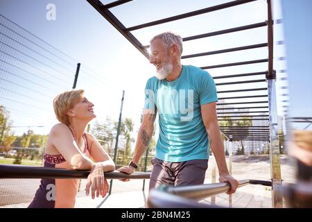 Heureux homme mature barbu dans une grande forme faisant des retouches sur des barres parallèles et parlant avec sa femme souriante. Couple senior actif et en bonne santé Banque D'Images