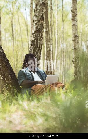 Homme africain joyeux qui fait du travail à distance sur l'ordinateur portable dans la nature. Photo verticale à faible angle Banque D'Images