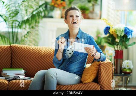 Bonne femme de ménage élégante en Jean chemise avec tablette PC étudier en ligne à la maison moderne en journée ensoleillée. Banque D'Images