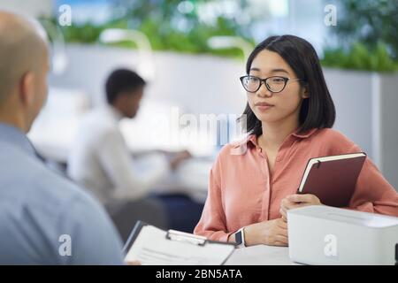 Portrait de la jeune femme d'affaires asiatique parlant au patron ou au Manager tout en se tenant dans l'intérieur moderne de bureau, espace de copie Banque D'Images