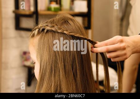 Processus de tressage le maître tisse des tresses sur sa tête blonde petite fille dans le salon de beauté gros plan. Soins professionnels des cheveux et création de coiffures. Banque D'Images