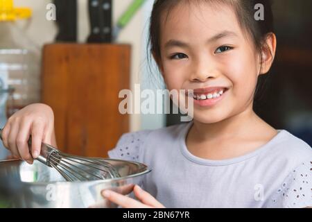 Portrait asiatique bonne fille, sourire mignon avec petite dent et profiter de la première formation de cuisine à la maison. Gros plan petite fille tenant un fouet en acier inoxydable Banque D'Images